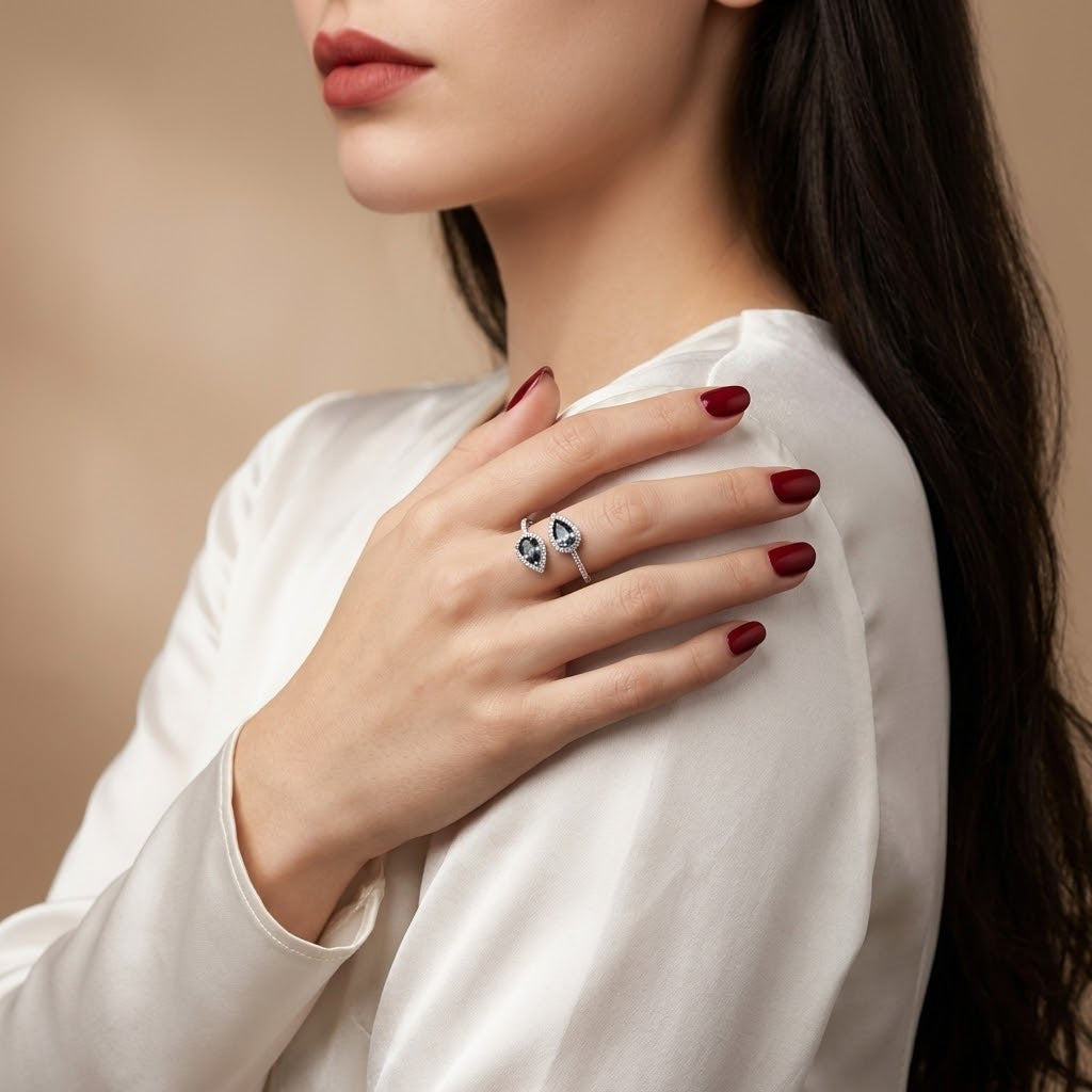  Woman wearing a silver ring with two small heart-shaped blue stones, with her hand resting on her shoulder. She is wearing a white satin top and has dark red nail polish.