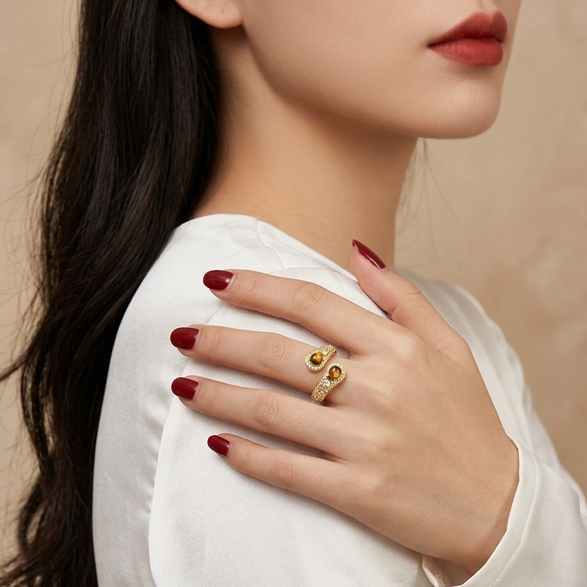 A close-up of a woman's hand wearing a gold ring with two oval-cut stones, styled with dark red nail polish and a white blouse against a soft beige background.