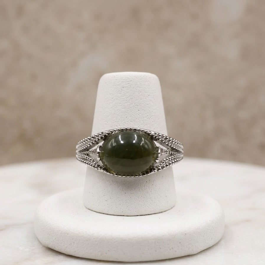 A silver ring with a round dark green cabochon gemstone, displayed on a white cone jewelry stand over a marble surface.