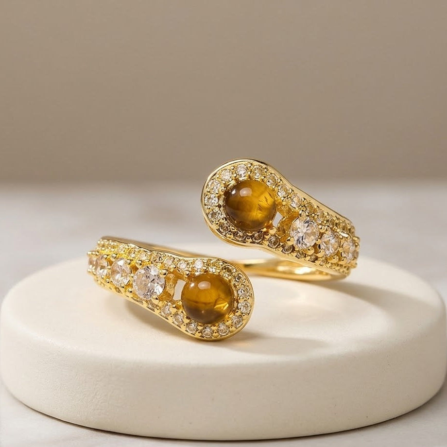 Two gold rings with oval brown gemstones and clear pavé accents, displayed on a round white ceramic riser against a soft beige background.