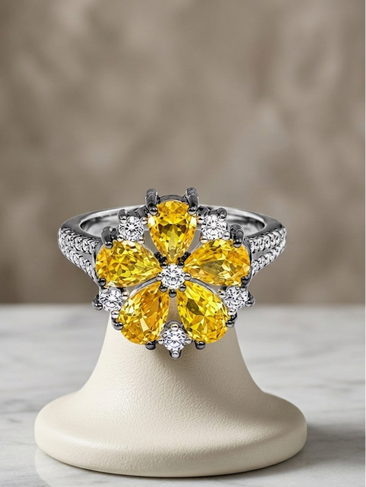 A floral cluster ring with large yellow gemstones and small clear stones on a silver-toned band, displayed on a cream jewelry stand over a marble surface against a soft beige background.