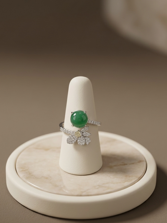 A silver ring with a round green gemstone and a floral cluster of small clear stones, displayed on a white cone ring stand over a round marble tray against a warm taupe background.