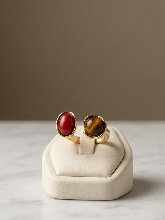 Two gold rings displayed on a cream ring box. One ring features a smooth, oval red cabochon stone, and the other features an oval brown and black tiger's eye stone, set against a neutral background.