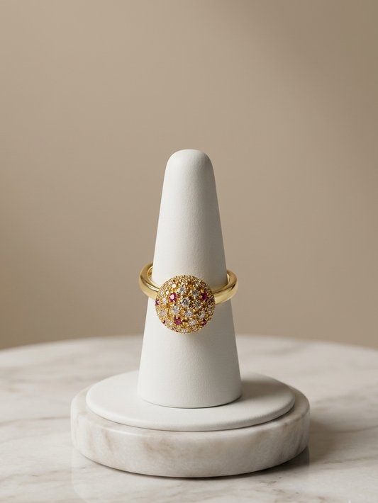 A gold ring with a round, textured multicolor stone sits on a white cone ring holder placed on a white marble surface against a warm beige background.