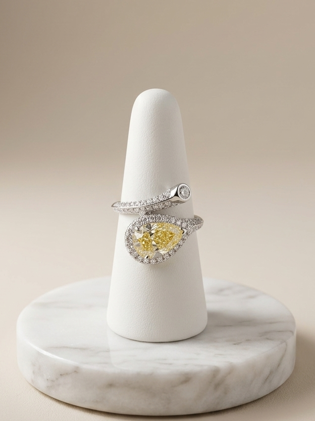 A silver ring with a heart-shaped yellow gemstone surrounded by small clear stones, displayed on a white cone stand atop a round marble base against a warm beige background.