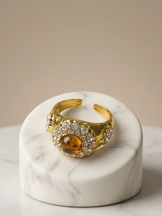 A gold-toned open cuff ring with a central amber oval stone and surrounding clear pavé stones, displayed on a white marble cylindrical riser against a warm beige background.