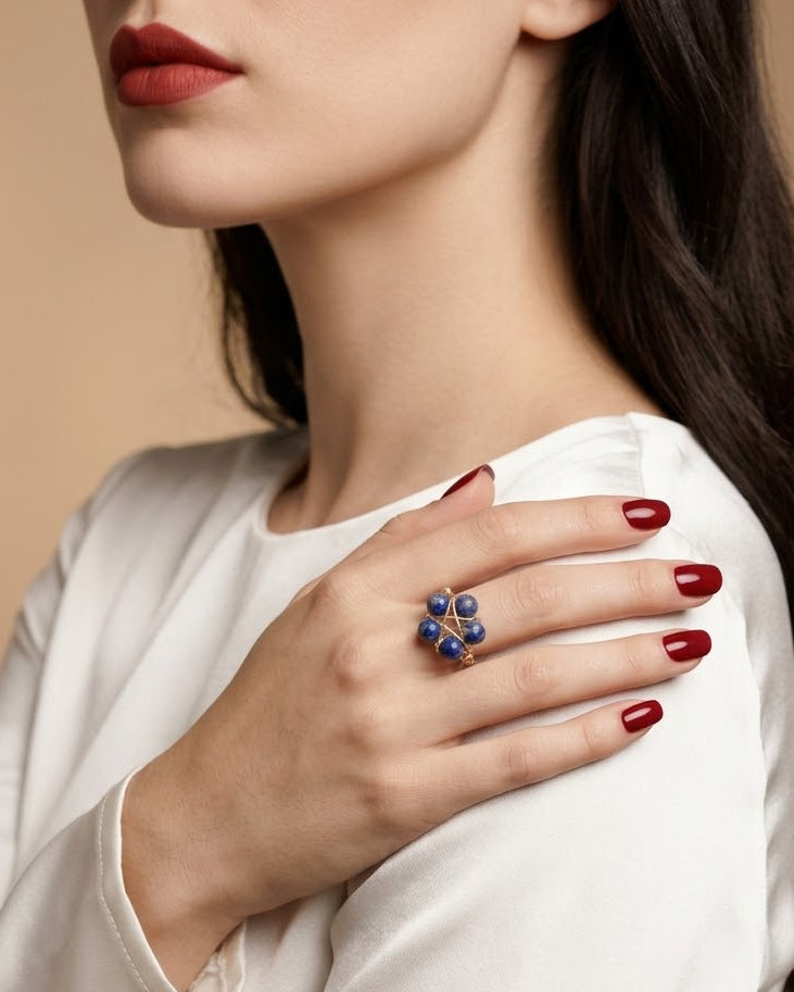 A close-up lifestyle shot of a model wearing a distinctive ring with blue and white stones on her ring finger, paired with a white satin top and dark red nail polish against a warm beige background.