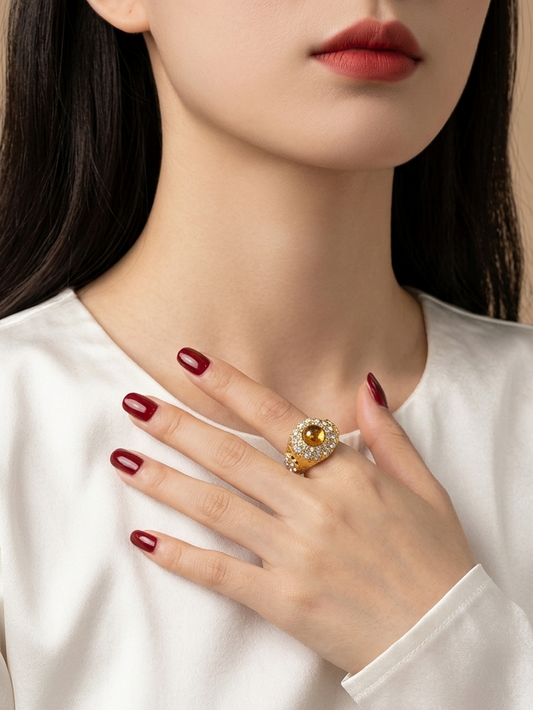 A close-up of a woman wearing a large gold ring with a yellow gemstone and clear accent stones on her finger, paired with a white satin top and dark red nail polish against a warm beige background.