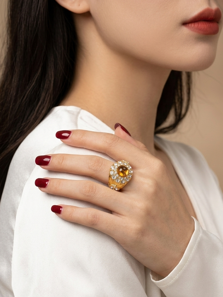 A close-up of a woman wearing a large yellow gemstone ring with a halo setting on her hand, which is resting on her shoulder. She is wearing a white satin top, and the background is a soft beige studio backdrop.