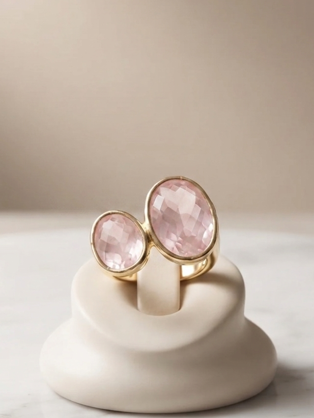 Two gold rings with oval pink gemstones displayed on a small cream-colored stand on a marble surface against a soft beige background.