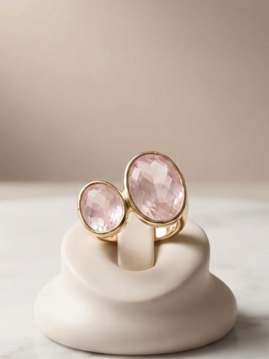 Two gold rings with oval pink gemstones displayed on a small cream-colored stand on a marble surface against a soft beige background.