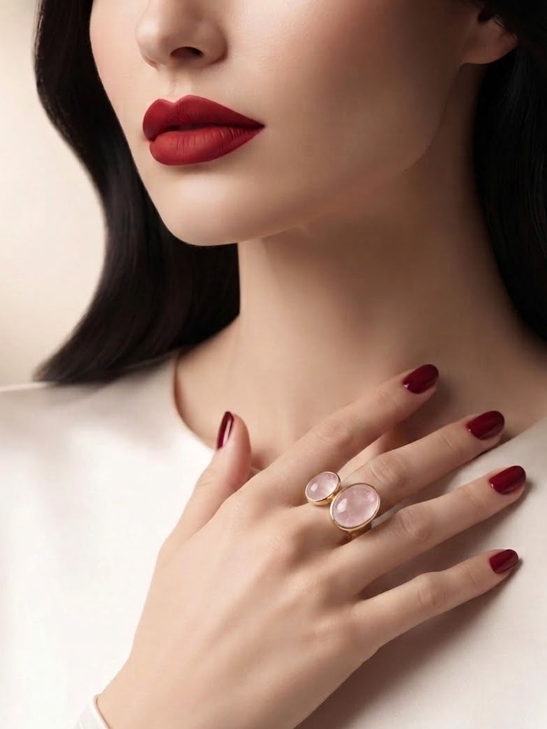 A close-up of a woman wearing a gold ring with two oval pale pink gemstones, styled with deep red lipstick and matching red nail polish.