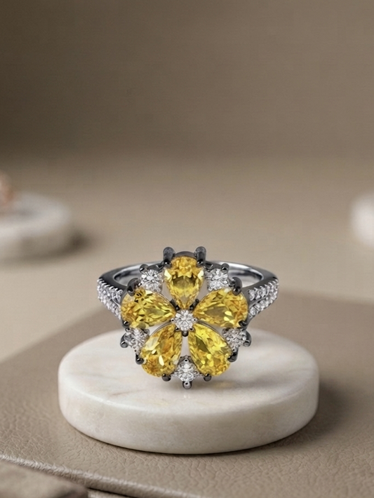 A floral cluster ring featuring large yellow gemstones and small clear stones on a silver-toned band, displayed on a round white marble stand against a warm beige background.