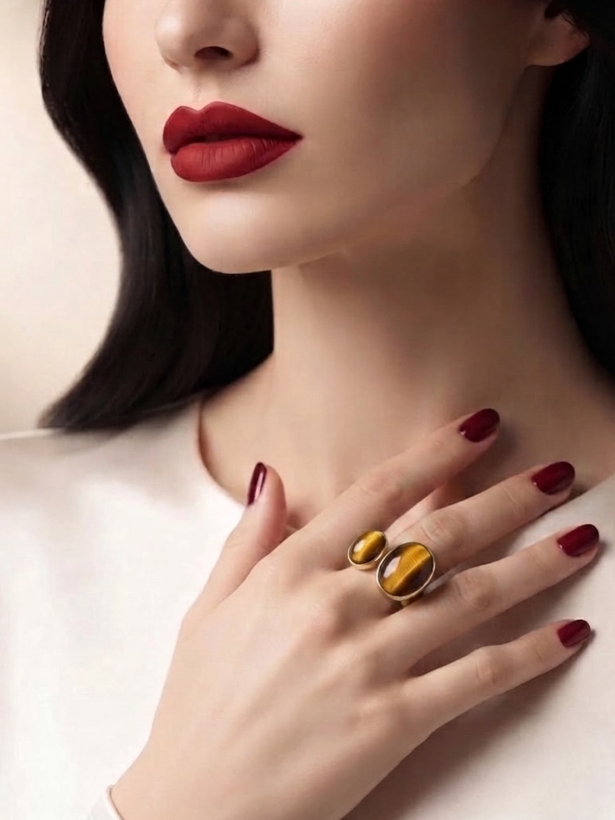 A close-up of a model's hand and lower face wearing a statement ring with two oval tiger's eye stones in a gold setting, against a soft beige background.