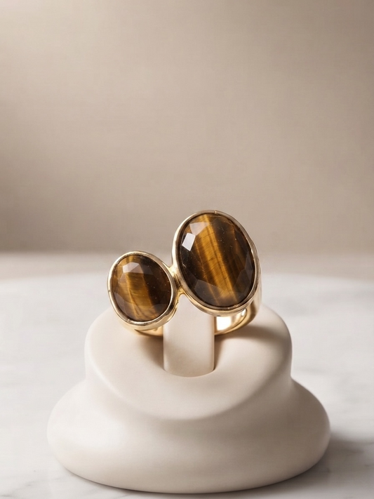 Two gold rings with large oval tiger's eye stones, displayed on a white ceramic ring stand against a neutral beige background.