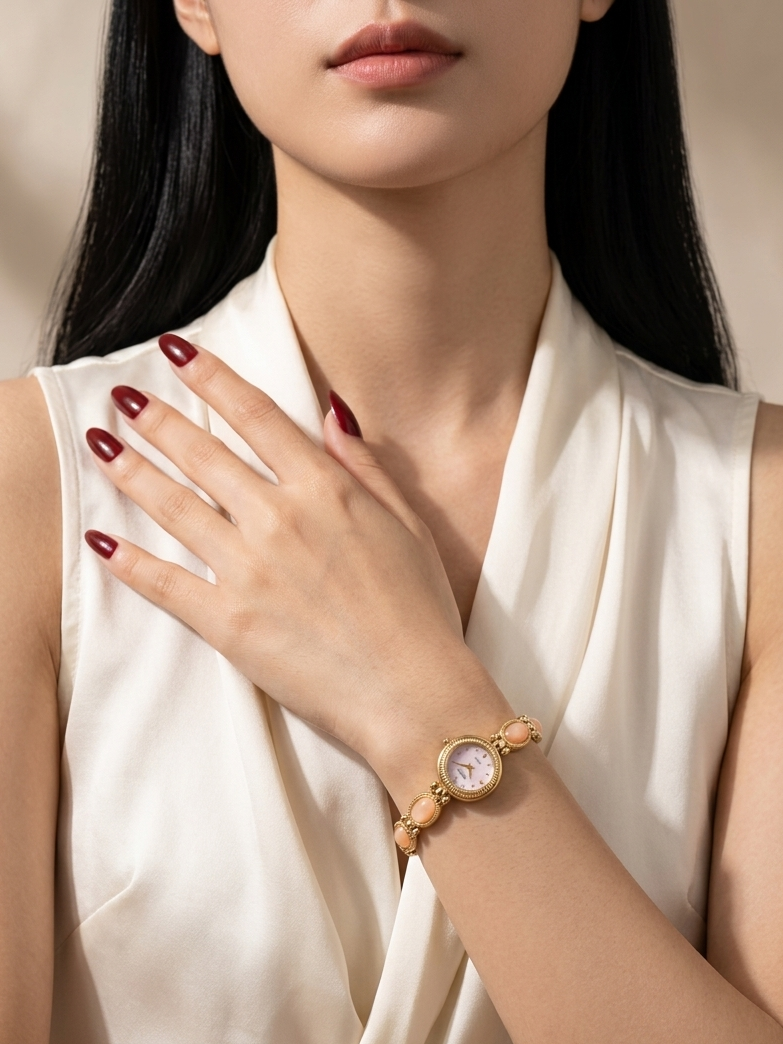 A close-up of a woman wearing a gold-toned watch with a round dial and matching bracelet, paired with a cream satin sleeveless top. Her hand is resting on her chest, showcasing the watch on her wrist.
