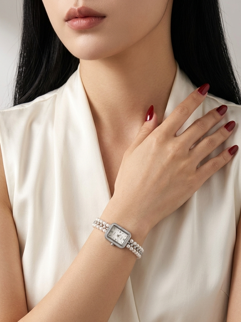 A woman wearing a silver-toned rectangular watch with a woven strap, paired with a cream satin sleeveless top. Her hand with dark red nail polish rests near her collarbone.
