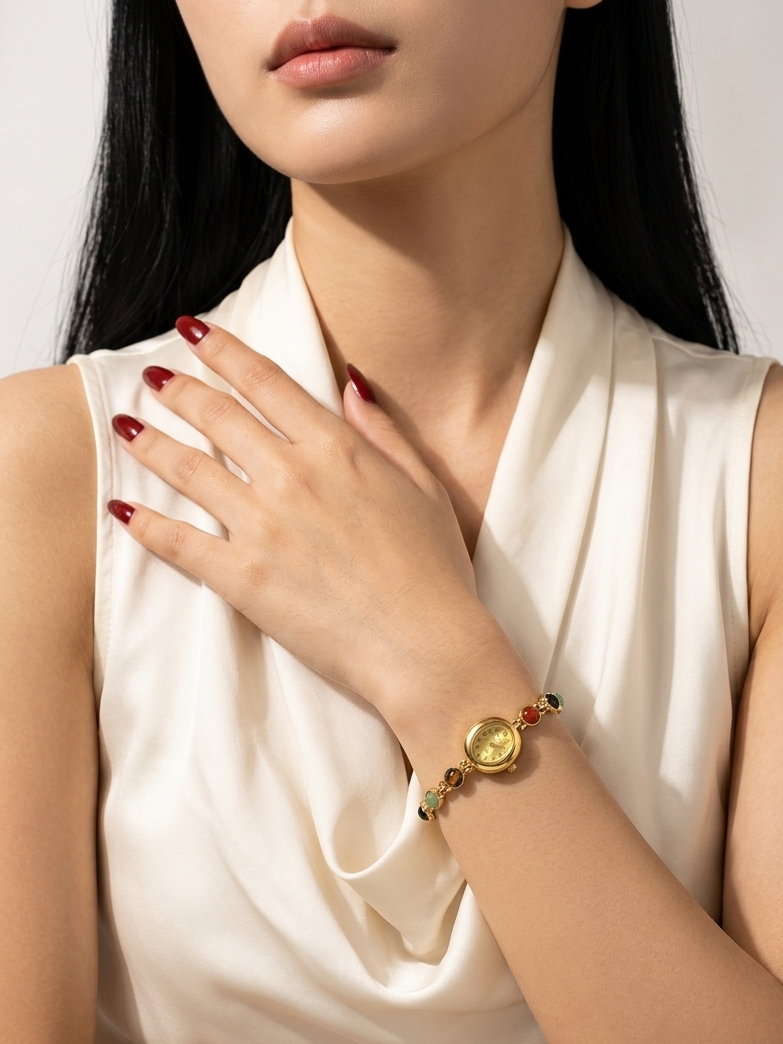 A close-up of a woman wearing a gold bracelet with green and red details, paired with a cream-colored satin top. The image highlights the bracelet on her wrist.