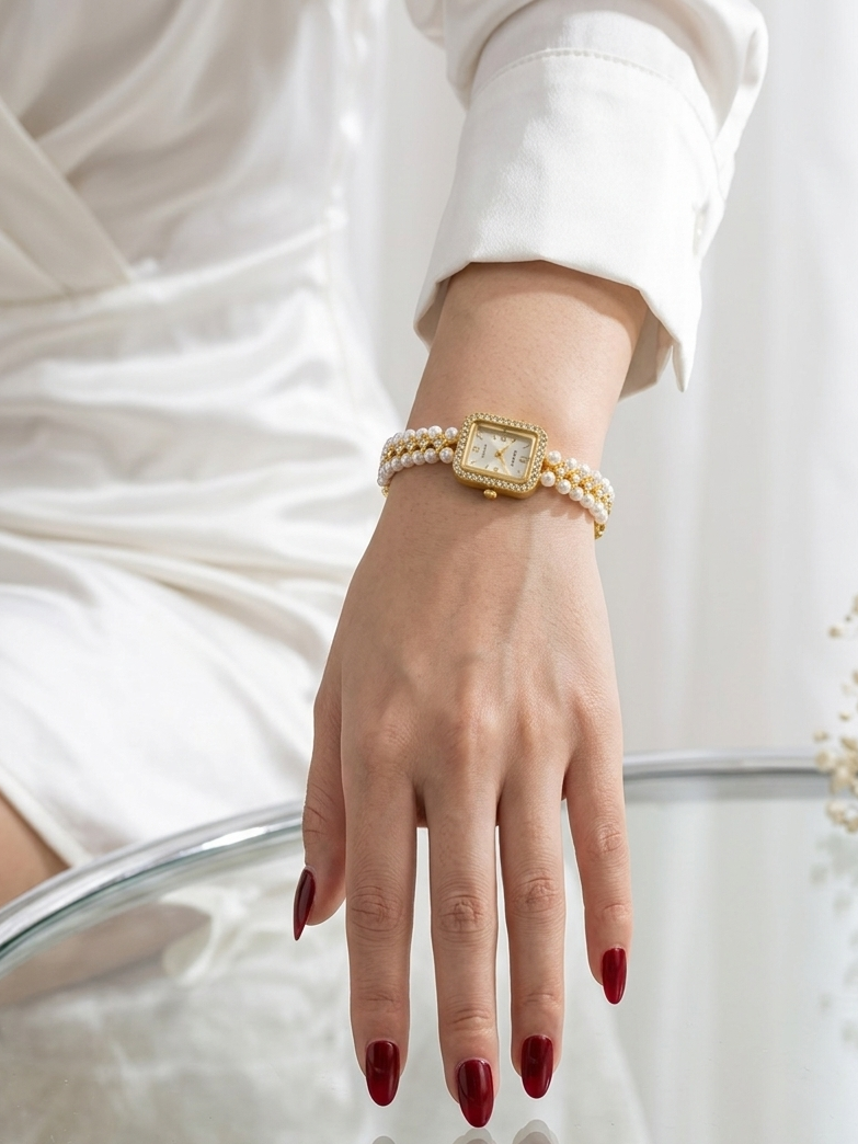 A close-up of a woman's hand with dark red manicured nails resting on a glass table, wearing a gold watch with a rectangular face and a double-strand pearl bracelet.
