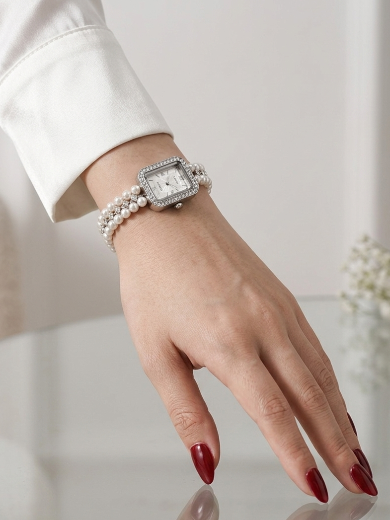 A woman's hand with dark red manicured nails resting on a glass table, wearing a rectangular watch with a diamond-encrusted bezel, white face, and a double-strand pearl bracelet.