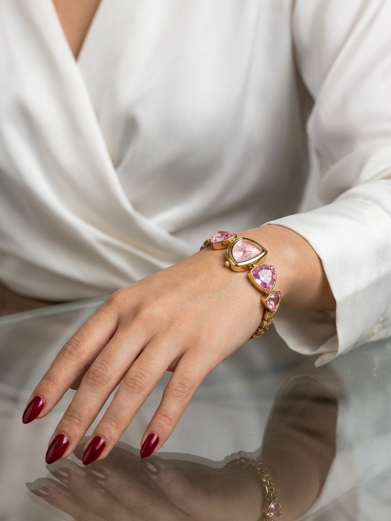 A gold bracelet with three pink heart-shaped gemstones worn on a woman's wrist, shown in a lifestyle setting with a white satin blouse and a reflective glass table.