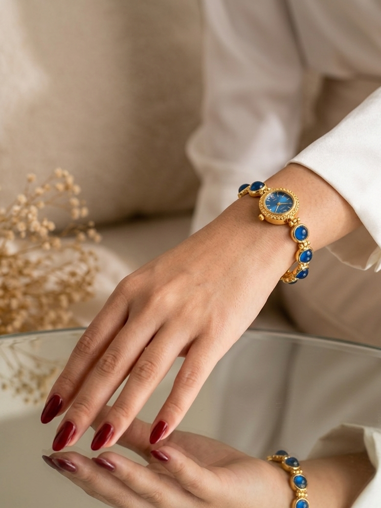 A close-up of a person's hands wearing a gold bracelet with round blue cabochon stones, paired with a white long-sleeve blouse. The image includes a clear glass table surface reflecting the hands and bracelet.