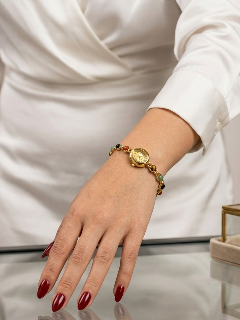 A gold-toned beaded bracelet with a round medallion and small green and red accents, worn on a woman's wrist. The woman is wearing a white satin dress, and the bracelet is shown resting on a reflective glass table.