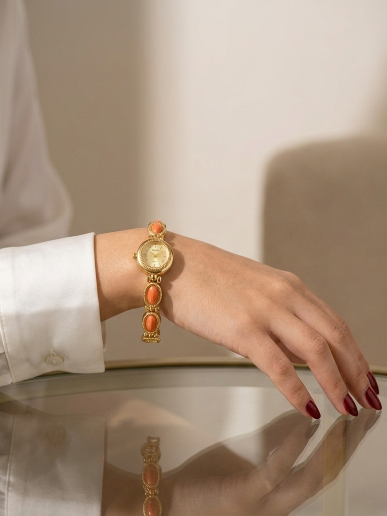 A gold-tone watch with orange stone accents worn on a woman's wrist, shown in a lifestyle setting with a white dress sleeve and a glass tabletop reflection.