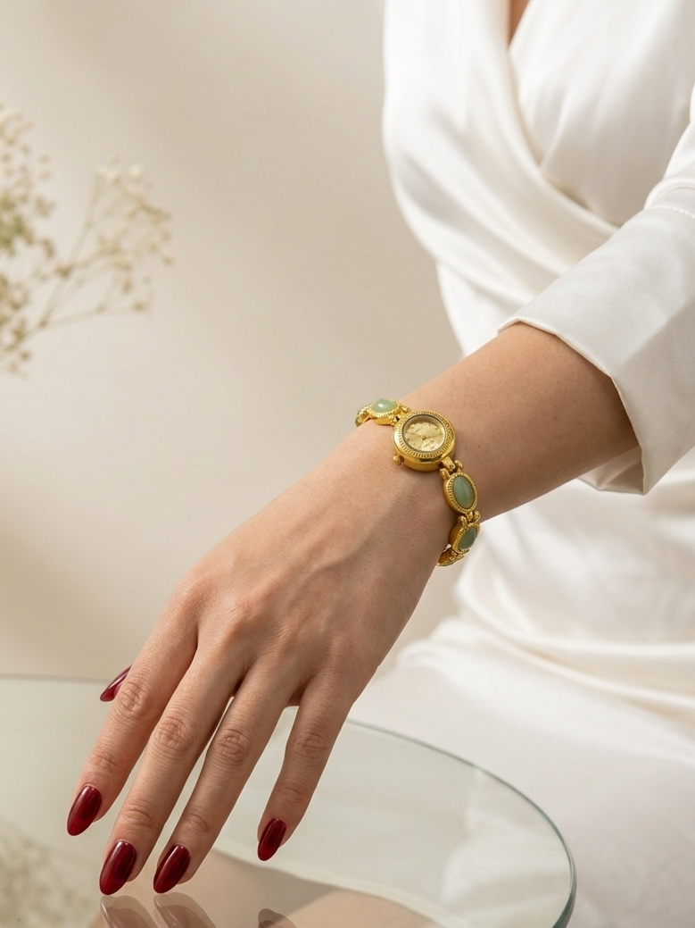 A gold-tone watch with oval green stone accents is worn on a woman's wrist, shown in a lifestyle setting with a white dress, red manicured nails, and a glass table.