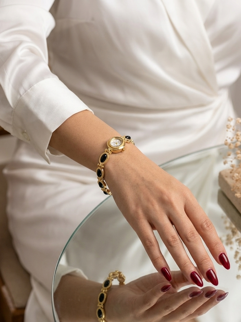 A gold bracelet with black oval inlays and a round white face, worn on a woman's wrist. The woman is wearing a white satin dress and resting her hand on a glass table, with the bracelet reflected in the glass.