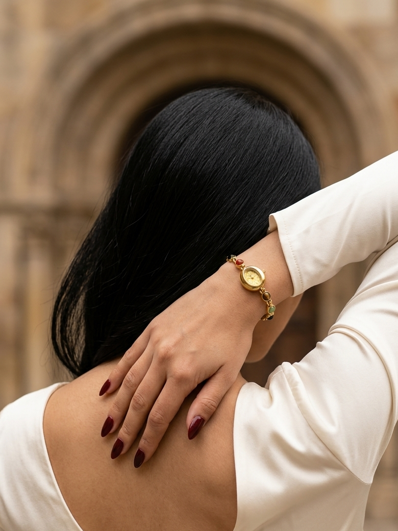 A woman with long black hair, seen from behind, wearing a gold watch on her wrist and a cream-colored long-sleeve top. She is posing in front of a blurred stone archway.