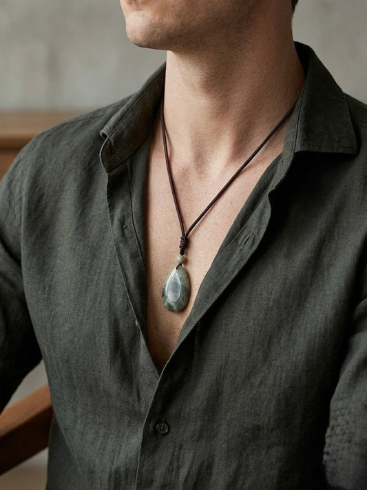 A close-up of a man wearing a dark charcoal linen shirt and a dark brown cord necklace with a polished gray teardrop pendant.