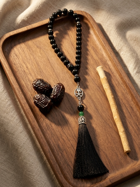 A black beaded tassel necklace with silver-toned and green accents, displayed on a wooden tray alongside three dates and a small wooden stick on a beige fabric background.