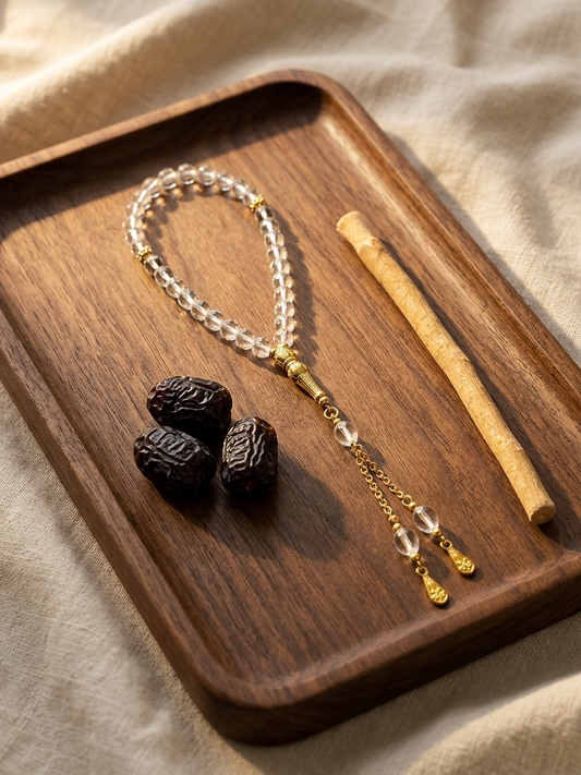 A beaded necklace with clear crystal-like round beads and gold-toned accents, displayed on a dark wooden tray beside three dark dates and a light wooden stick, set against a beige fabric background.
