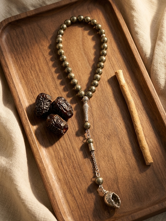 A beaded necklace with metallic bronze beads, a silver-toned central connector, and a dangling shell-shaped charm, arranged on a wooden tray with three dark dates and a wooden stick against a beige fabric background.