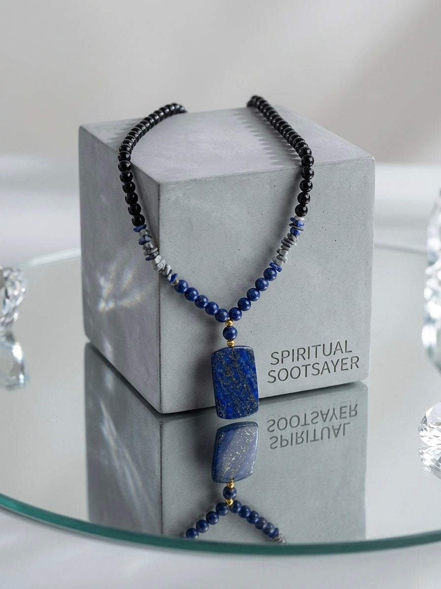 A beaded necklace with a rectangular blue stone pendant displayed on a gray cube stand. The necklace features dark beads, blue beads, and small metallic silver and gold accents.