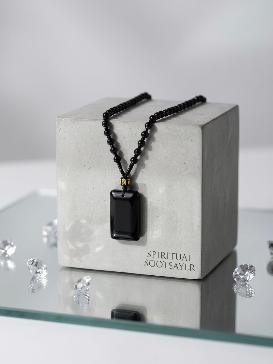 A black beaded necklace with a rectangular black pendant displayed on a light gray concrete block. The block has the text 'SPIRITUAL SOOTSAYER' printed on it.