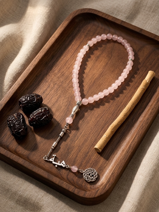 A pink beaded prayer rope with silver-toned metal accents and a calligraphy charm, arranged on a dark wooden tray with dates and a wooden stick on a beige fabric background.