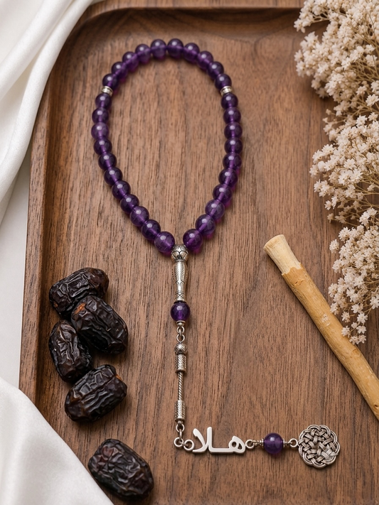 A beaded prayer rope (Tasbih) made of purple amethyst and silver-toned metal, displayed on a wooden tray with decorative dried flowers, dates, and a cinnamon stick.