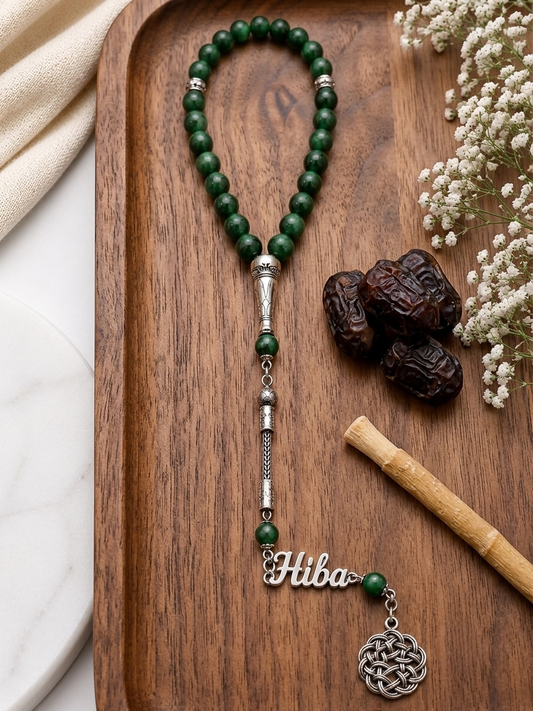 A beaded necklace with dark green polished beads, silver-toned spacer beads, a silver-toned nameplate reading 'Hiba', and a round silver-toned pendant, arranged on a wooden tray with decorative dried flowers, dates, and a bamboo stick.