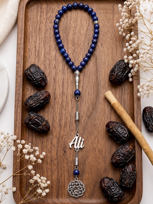 A beaded necklace with deep blue beads, silver spacer beads, a silver 'Ali' nameplate, and a silver Celtic knot pendant, displayed on a wooden tray with decorative dried flowers and dates.