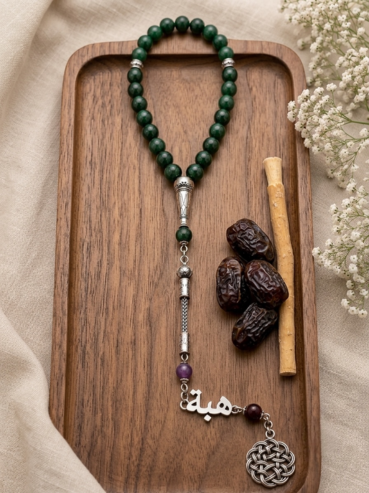 A beaded necklace with dark green round beads, silver-toned metal accents, a purple bead, and a silver Arabic calligraphy pendant on a wooden tray, styled with dates, a cinnamon stick, and white flowers.