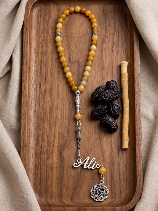 A beaded necklace with yellow marbled beads, silver-toned spacer beads, a silver nameplate charm reading 'Ali', and a round silver pendant, displayed on a wooden tray with dark dates and a cinnamon stick.