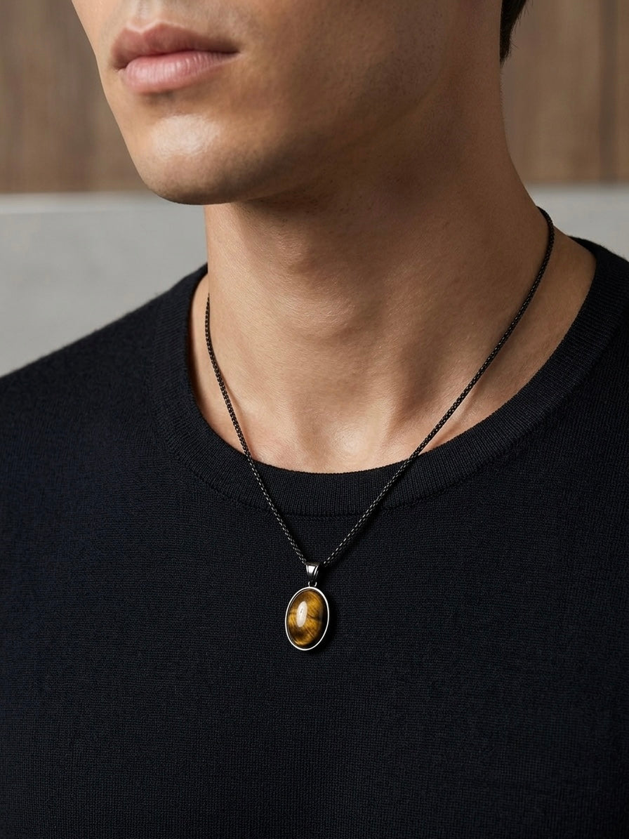 A close-up of a man wearing a black chain necklace with an oval pendant featuring a golden-brown tiger's eye stone.