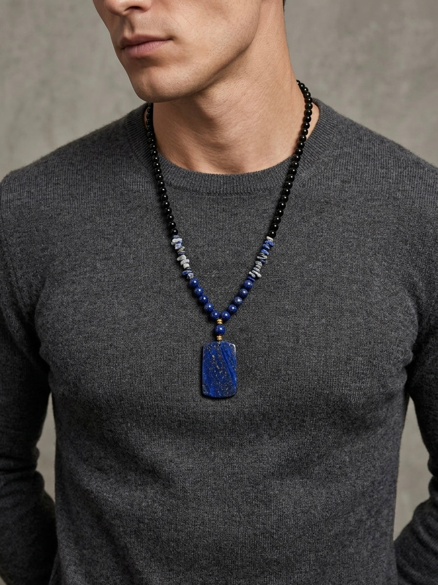 A man wearing a dark gray knit sweater and a beaded necklace with a rectangular blue pendant, shown from the chest up against a textured gray background.