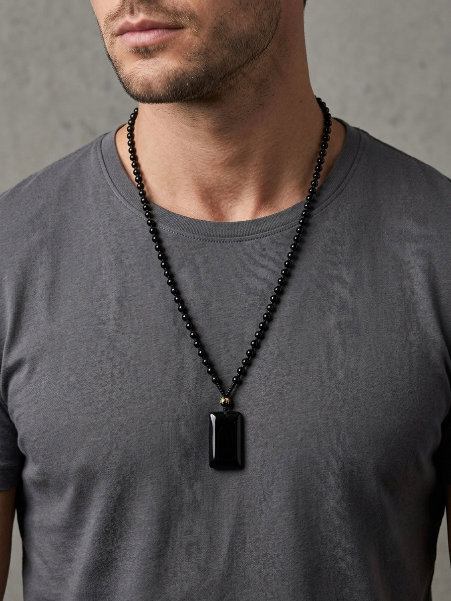 A man wearing a black beaded necklace with a rectangular black stone pendant, paired with a dark gray t-shirt against a textured gray wall.
