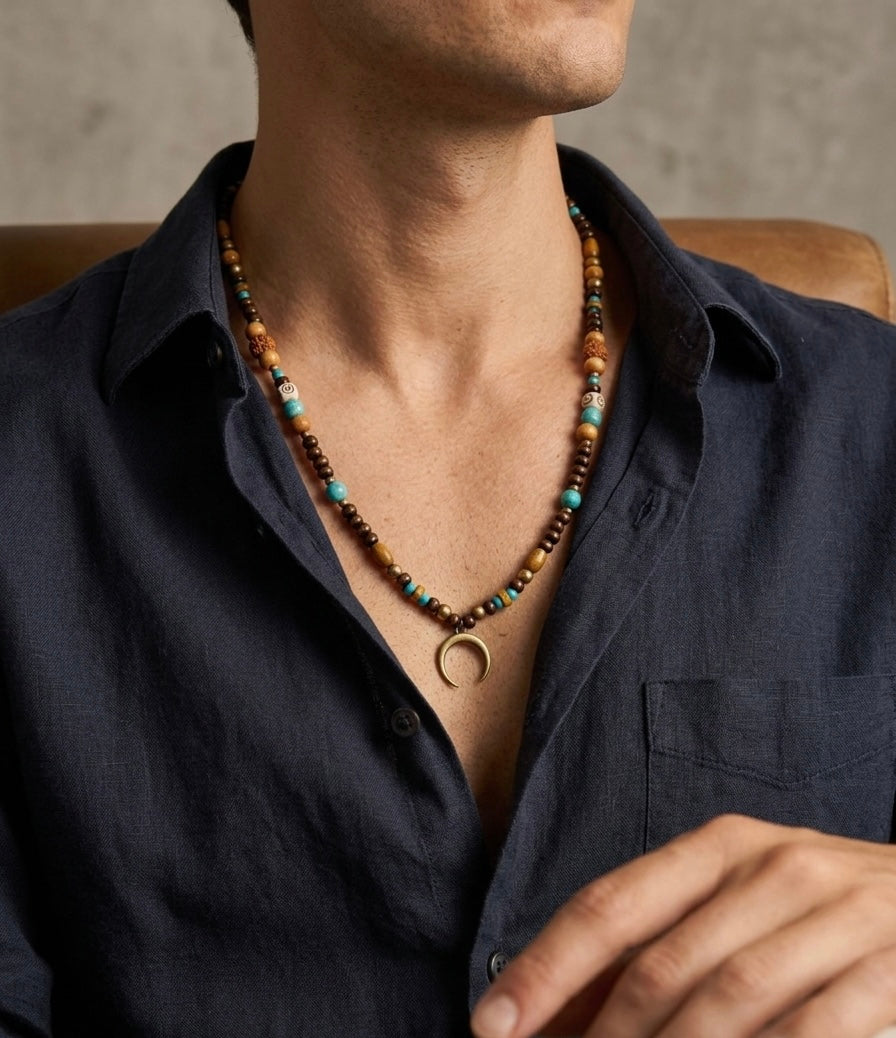 A close-up of a man wearing a dark navy blue linen shirt and a beaded necklace with turquoise, brown, and gold-colored beads, featuring a crescent moon pendant.