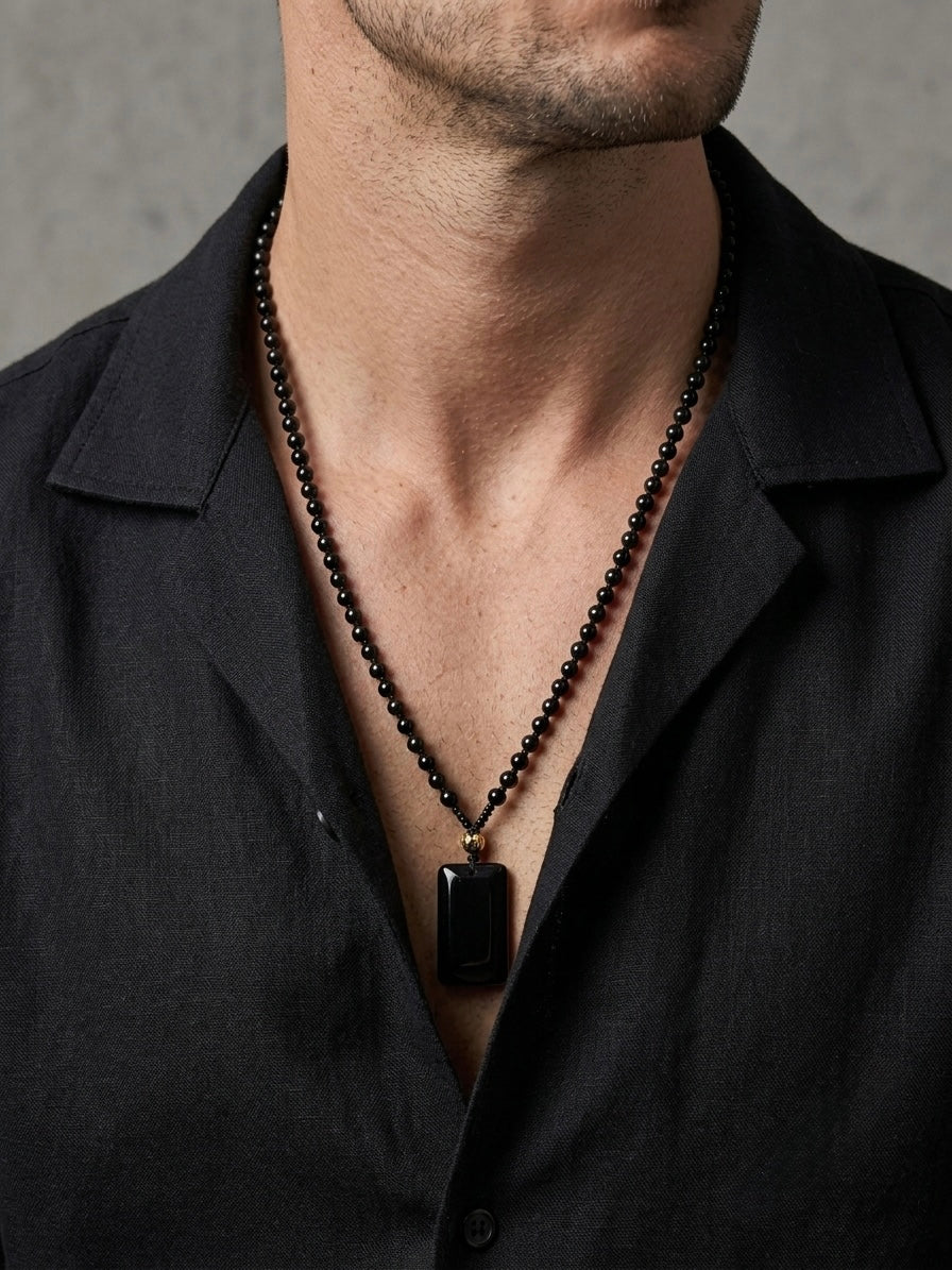 A close-up of a man wearing a black beaded necklace with a rectangular black pendant, styled with a black button-down shirt against a textured gray background.