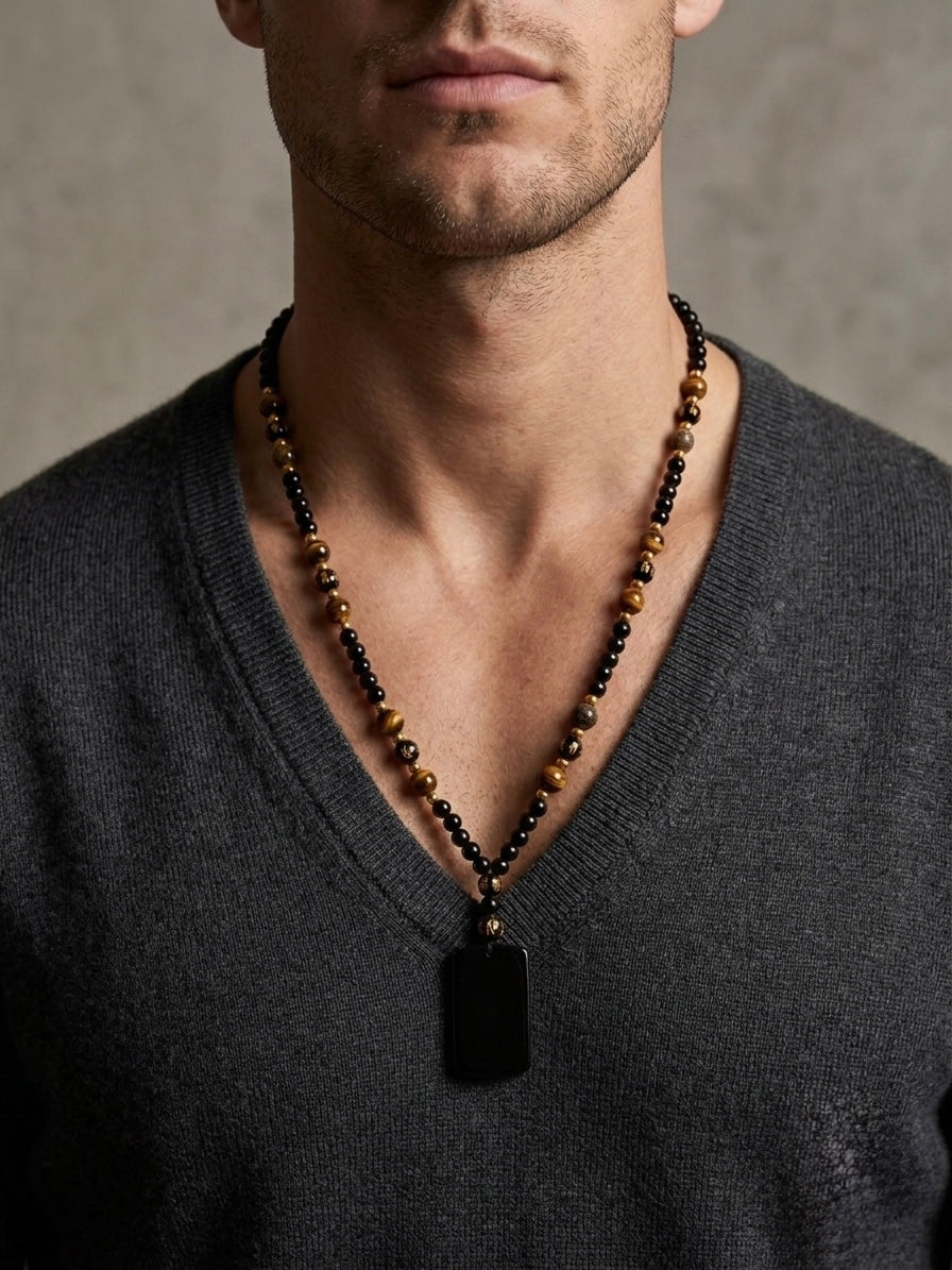 A close-up of a man wearing a beaded necklace with a black rectangular pendant over a dark gray V-neck sweater.