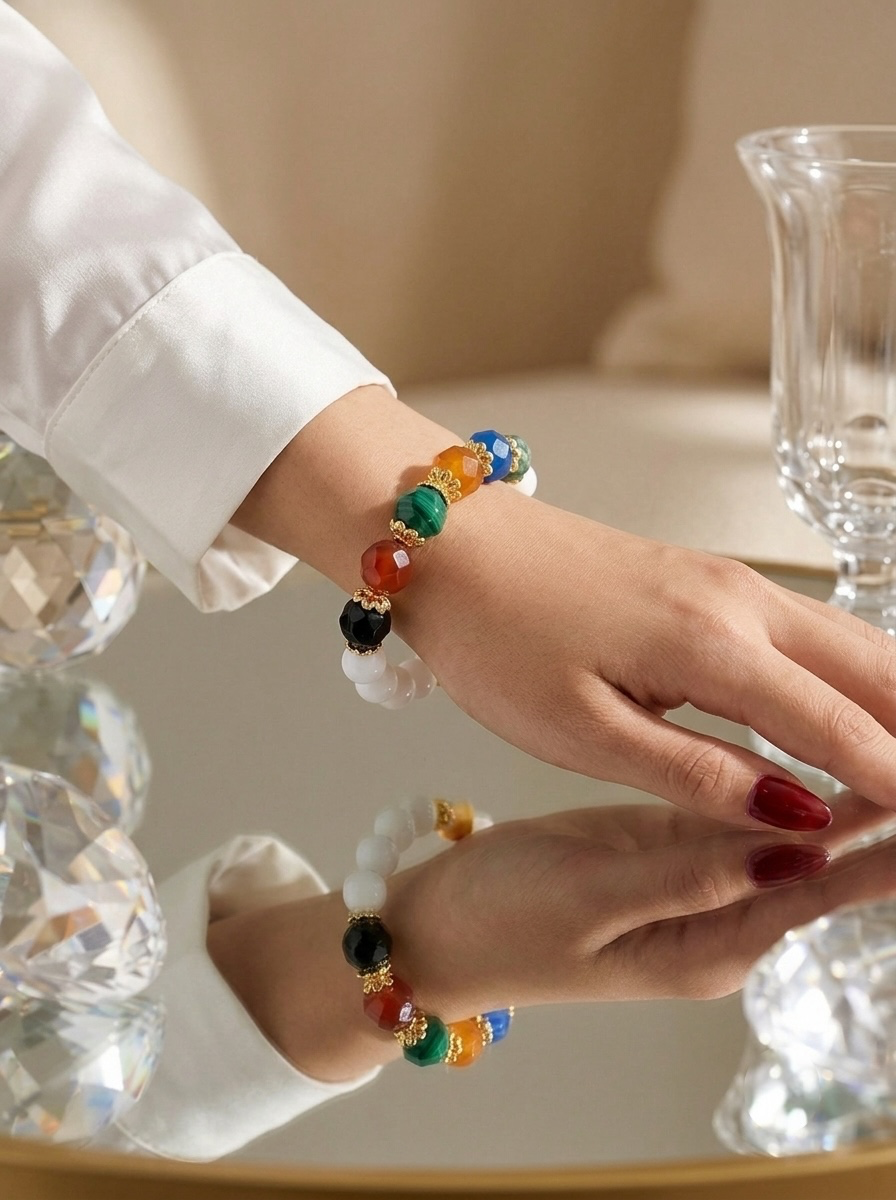 A close-up of a person's hand and wrist wearing a beaded bracelet with colorful round beads, gold-toned spacer details, and a white pearl-like bead, shown against a softly lit indoor background with a mirror reflection.